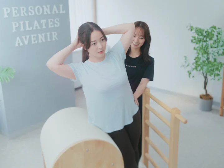 ラダーバレルの前で身体を伸ばす女性と、うしろで見守るトレーナー
